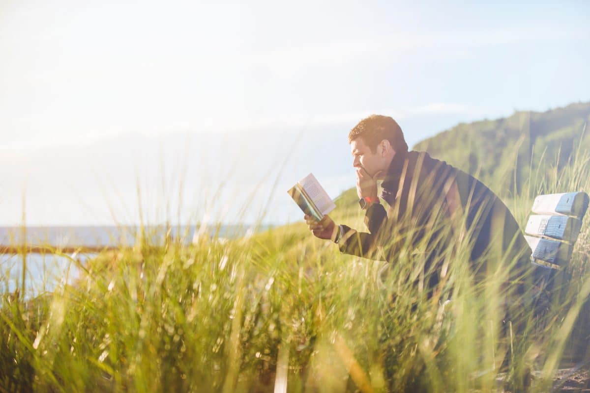 A man is reading a book outside while sitting on a chair. The grass around him is tall.