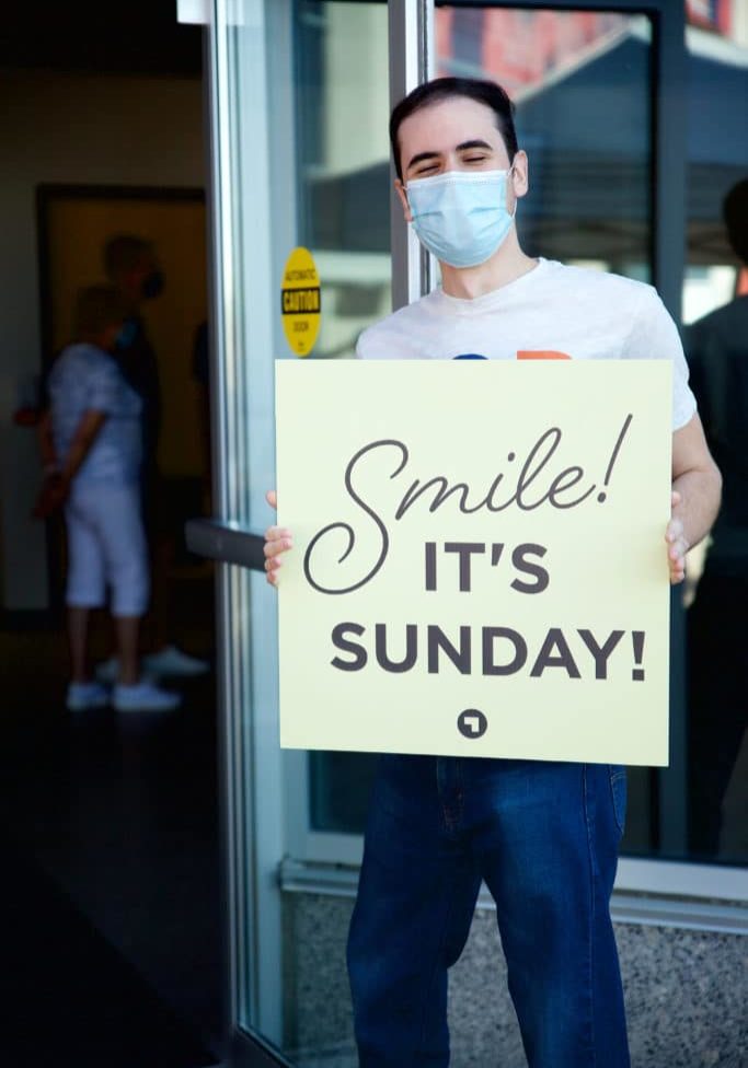 A man holds a sign outside of Connexus Church. The sign reads "Smile! It's Sunday!"